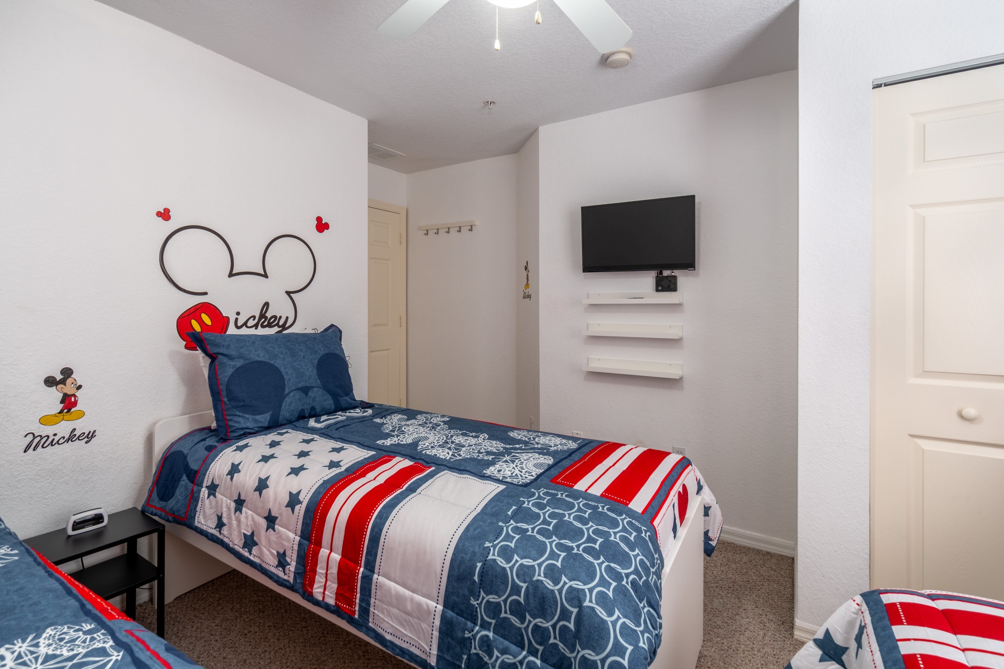 A bedroom with a Mickey Mouse wall decal, a bed with a star-patterned blanket, a TV mounted on the wall, and a door.