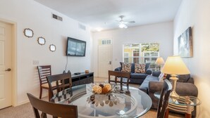 A living room with a glass-top dining table, a sofa, a TV mounted on the wall, and a ceiling fan. - Villas at Seven Dwarfs 4601 B8.106 (Kissimmee)