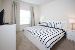 A bedroom with a large bed, a desk, a chair, a lamp, and a window with curtains.