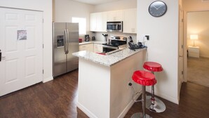 A modern kitchen with white cabinets, stainless steel appliances, a granite countertop, and red bar stools. - Lucaya Village 2905 - 101 (Kissimmee)
