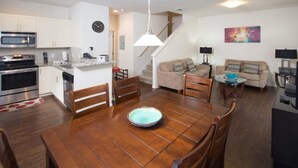 A modern kitchen with white cabinets, a microwave, and a stove. There is a dining table with chairs and a bowl on it. A living area with a sofa and a coffee table is visible. - Lucaya Village 2905 - 101 (Kissimmee)