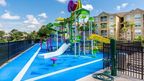 A colorful playground with slides and climbing structures, surrounded by a fenced area and residential buildings in the background. - Windsor Hills 2583 (Kissimmee)