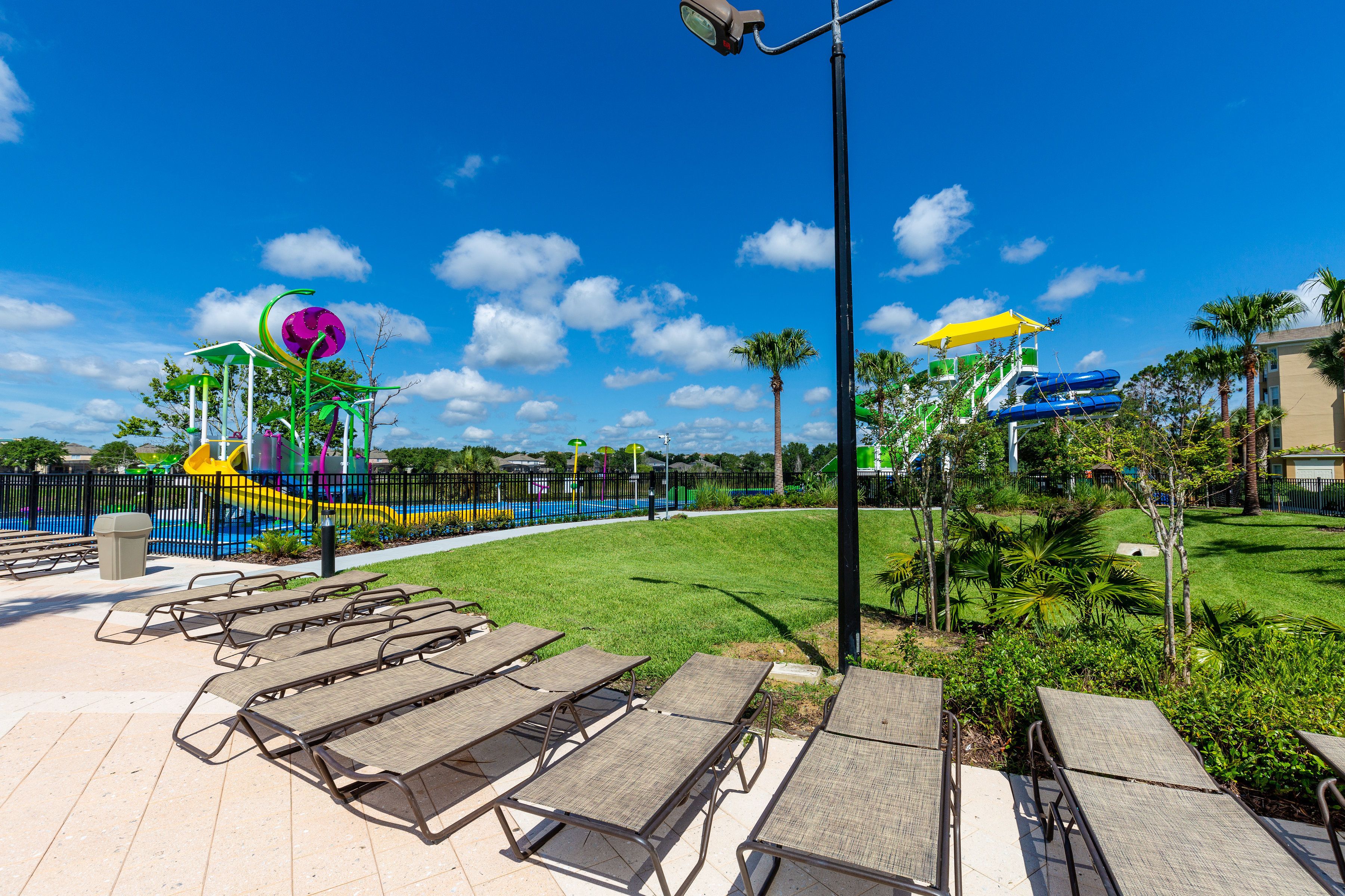 Zona exterior ensolellada amb un parc infantil, palmeres i seients de relax.