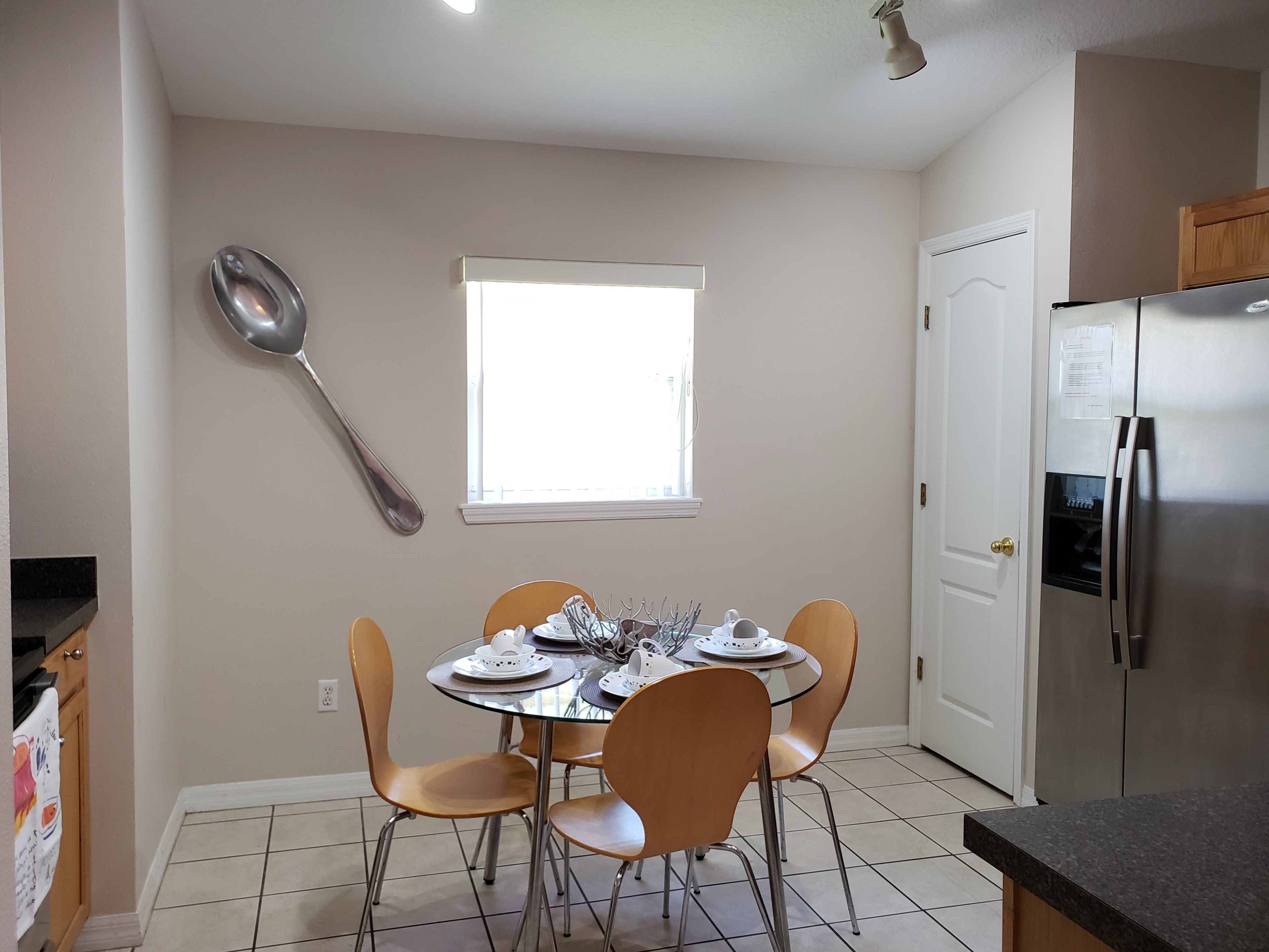A kitchen with a dining table, chairs, a spoon on the wall, a window, and a refrigerator.