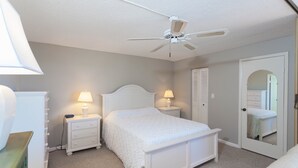 Une chambre à coucher avec un lit blanc, deux tables de chevet avec des lampes, un ventilateur de plafond, une porte avec un miroir et une commode verte.