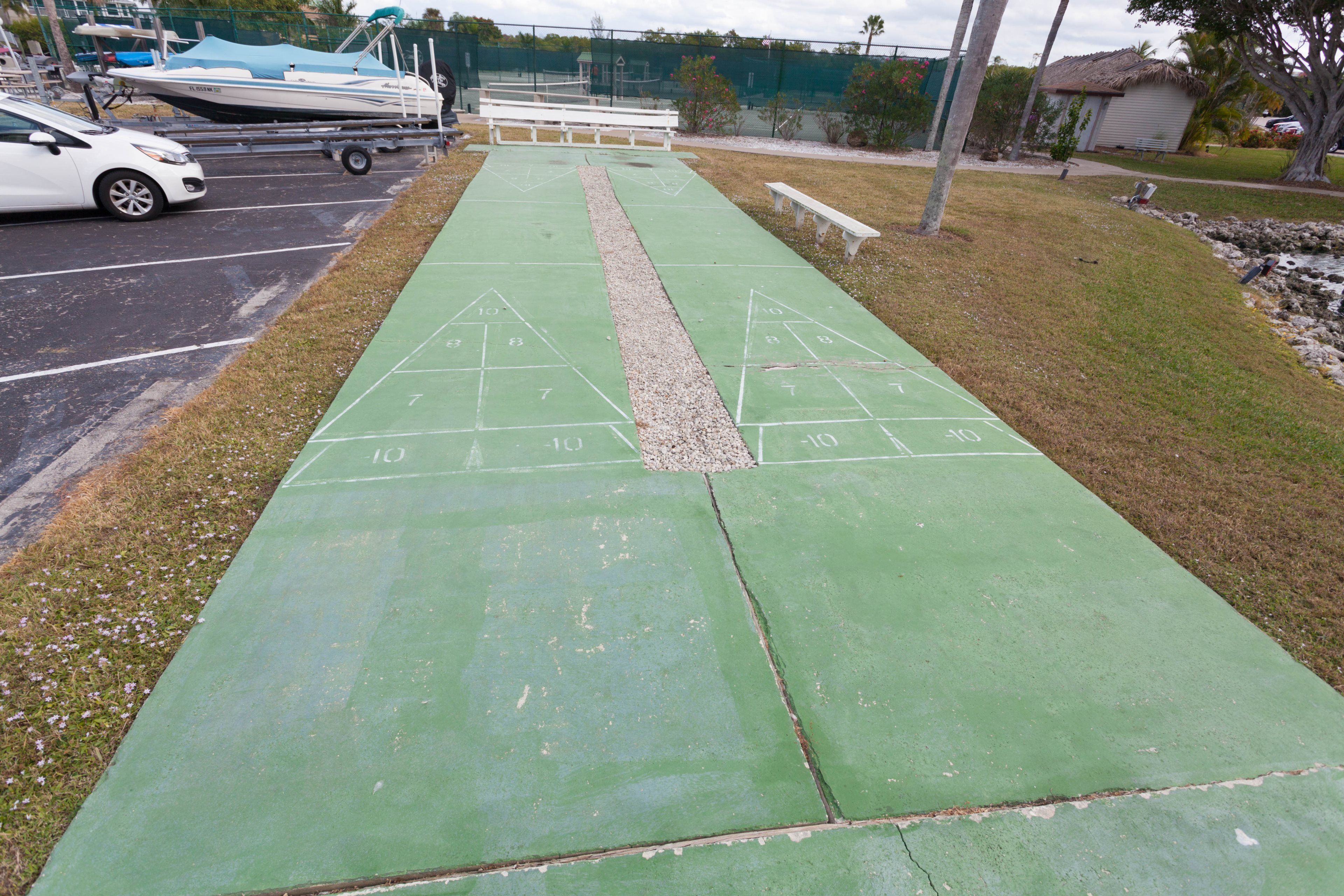 Un terrain de jeu de shuffleboard vert, avec des zones de marquage numérotées, une voiture blanche garée à proximité et une remorque pour bateau en arrière-plan.