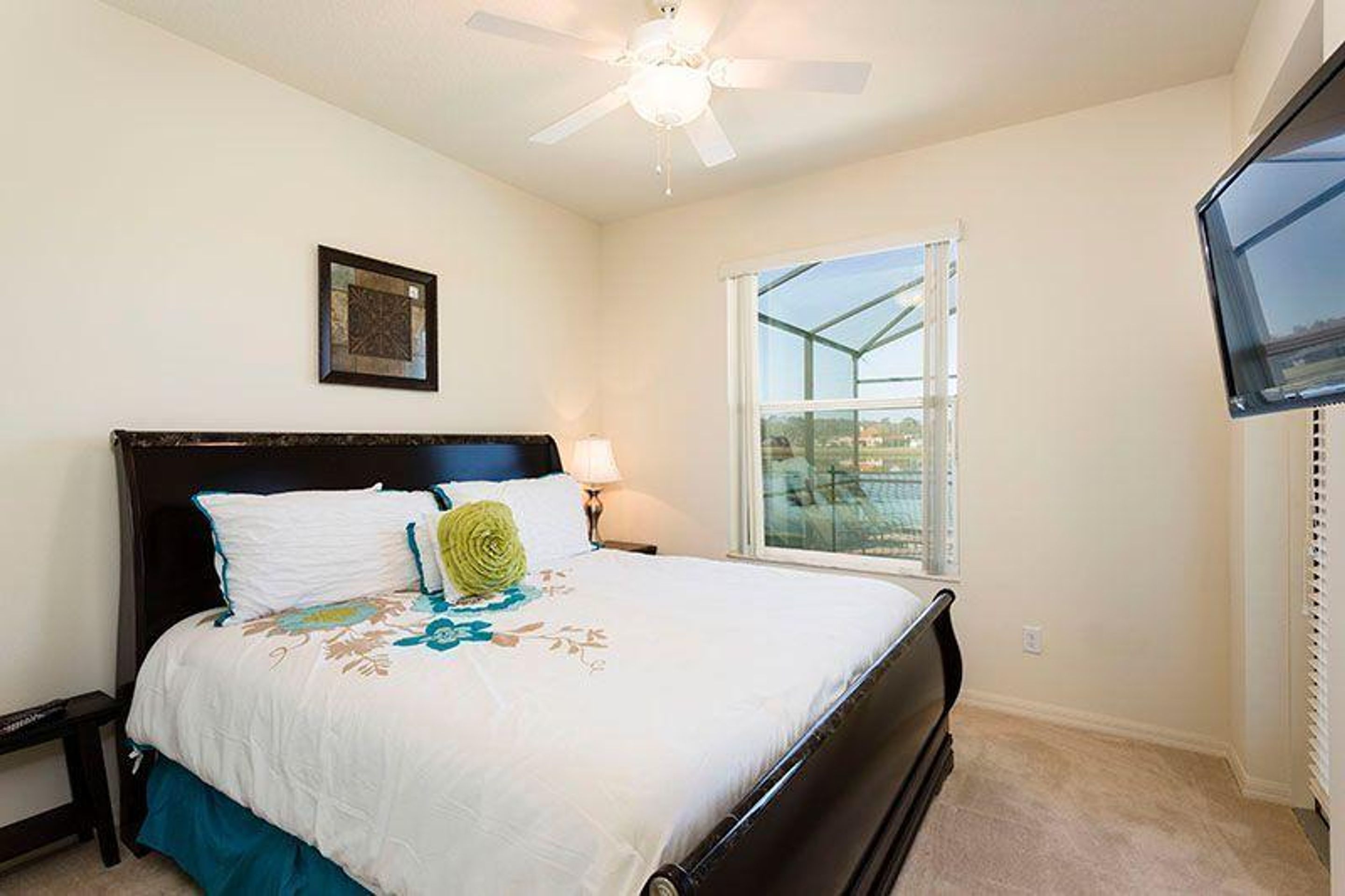 A bedroom with a bed, a ceiling fan, a window with a view, and a TV on the wall.
