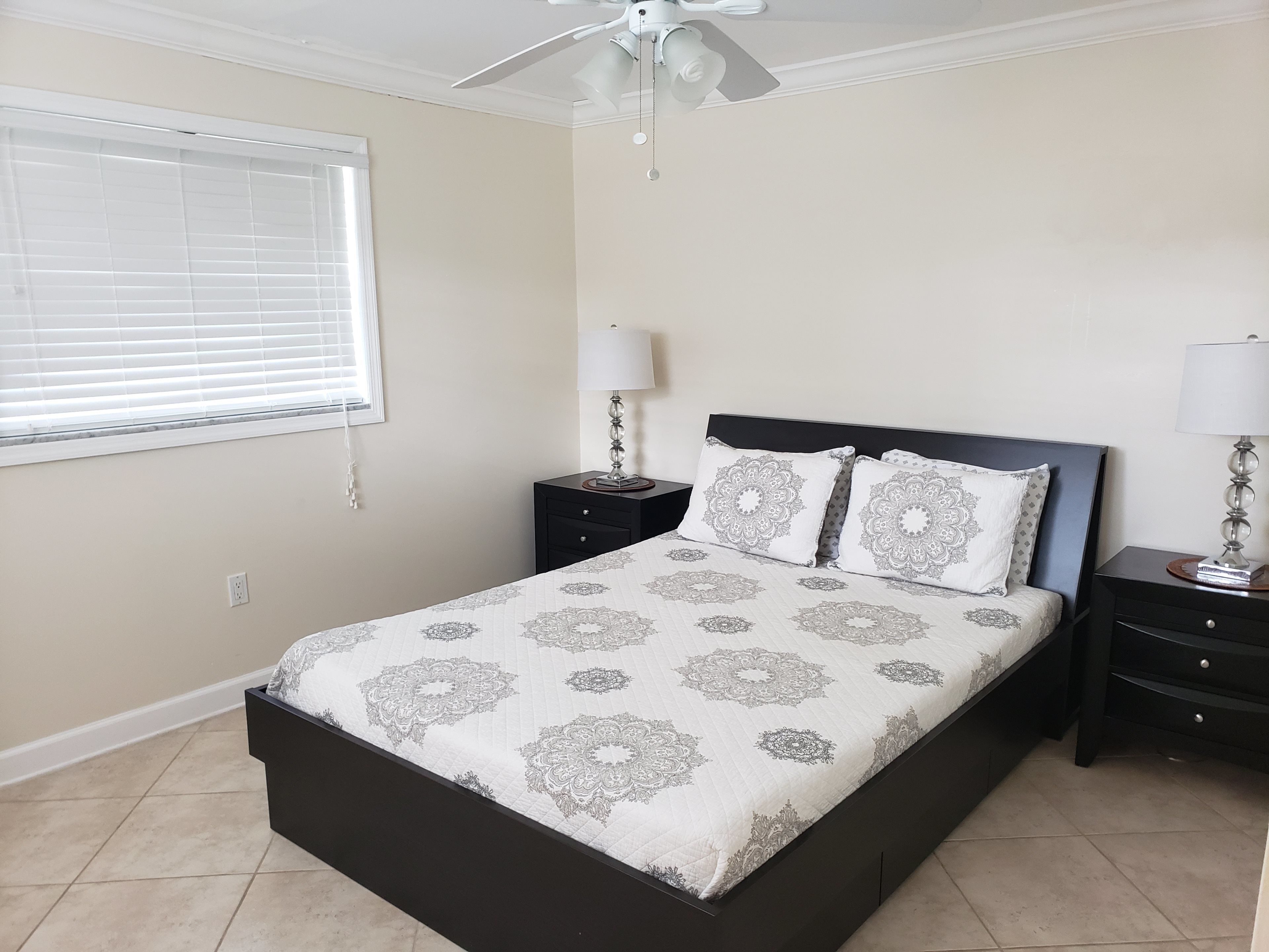 A bedroom with a bed, two bedside tables, a ceiling fan, a window with blinds, and a wall socket.