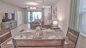 Ein Essbereich mit einem Holztisch, gedeckt für vier Personen, einem Fernseher im Hintergrund und einem Wohnzimmer mit einer Couch und einer Lampe.