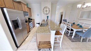 A modern kitchen with stainless steel appliances, a central island, and a dining area with white chairs and a wooden table. - Paradise Palms 3073 (Kissimmee)