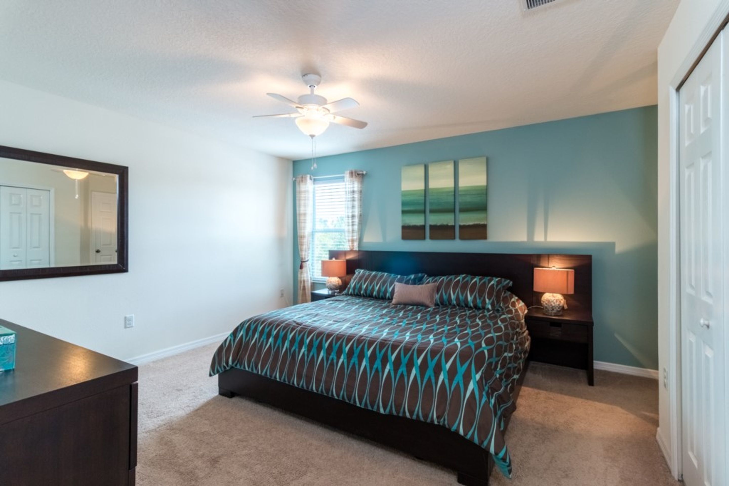 A bedroom with a bed, a ceiling fan, a nightstand with a lamp, a dresser, and a mirror on the wall.