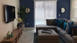 En stue med et fladskærms TV, en potteplante, en sofa med puder og et vindue med persienner.