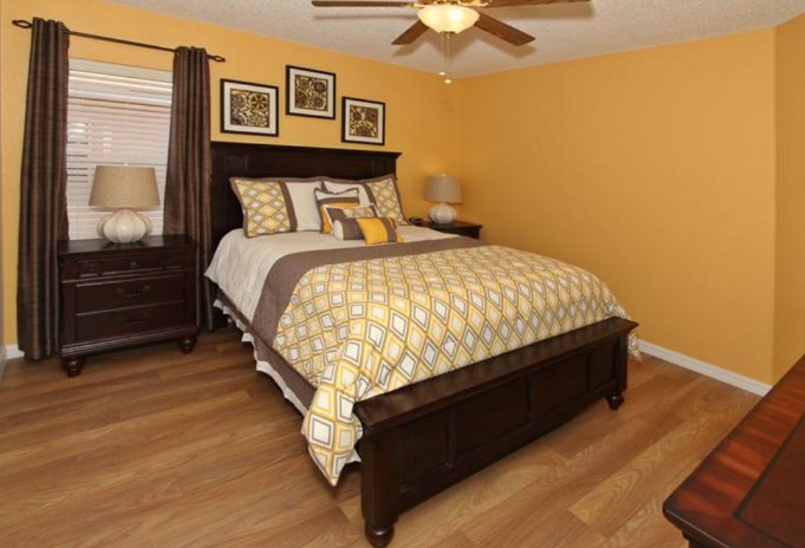 A bedroom with a wooden bed, two bedside tables with lamps, a ceiling fan, and three framed pictures on the wall.