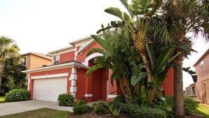 Una casa moderna de dos pisos con fachada roja, puerta de garaje blanca y un jardín delantero bien cuidado con una palmera de plátano grande y varios arbustos.