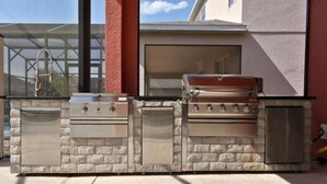 Una cocina exterior con una parrilla empotrada, horno y pileta, frente a una casa con un ventanal grande y puerta de vidrio.
