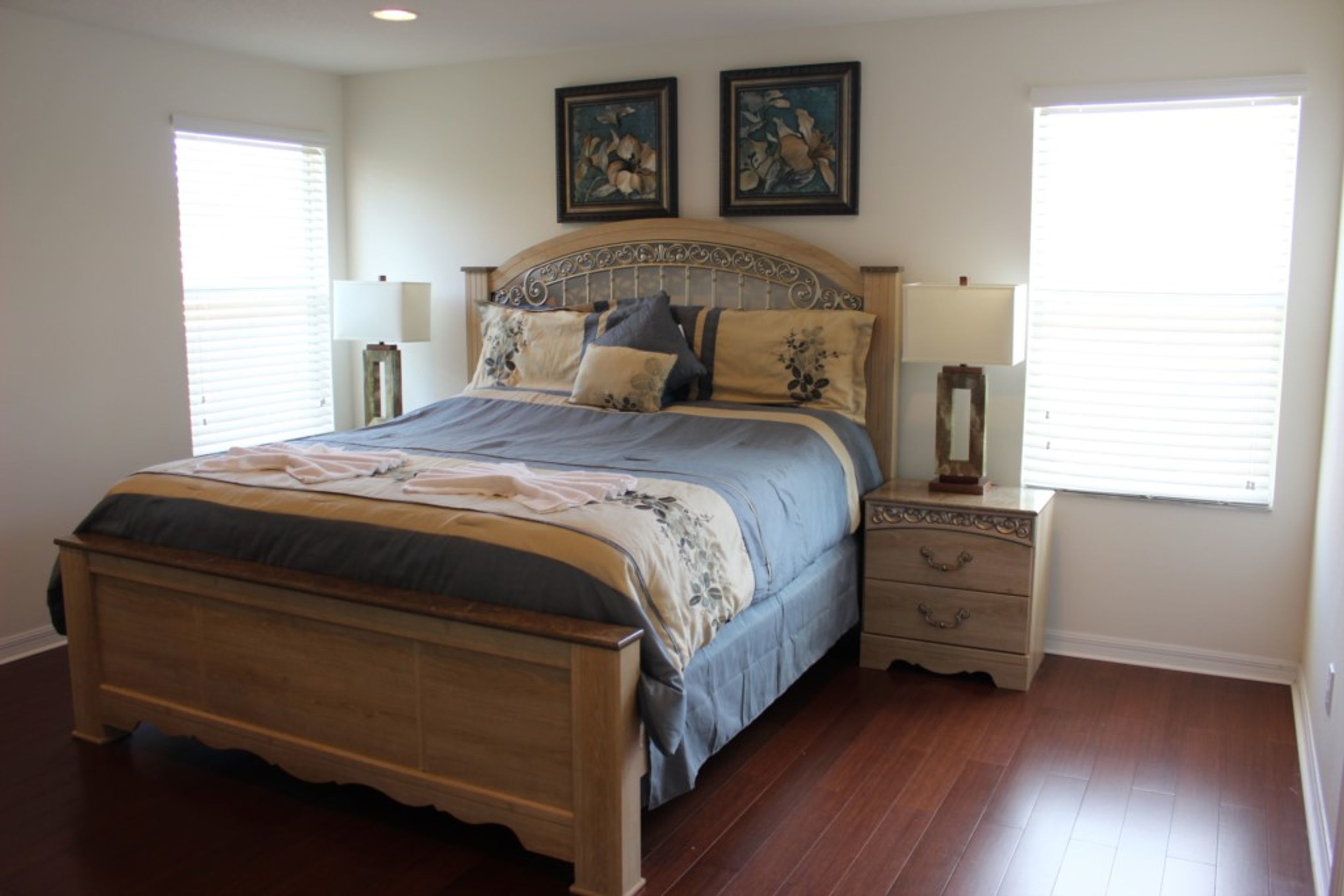 A bedroom with a wooden bed, two bedside tables, and a nightstand. There are two windows with blinds, and the walls are painted white.