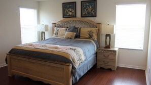 A bedroom with a wooden bed, two bedside tables, and a nightstand. There are two windows with blinds, and the walls are painted white.