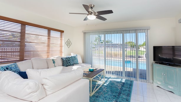 A living room with a white sofa, a ceiling fan, a TV on a cabinet, and a view of a pool through large windows. - Fort Myers Beach Area 540 (Fort Myers)