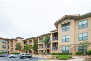 A multi-story residential building with balconies, a parking lot, and a landscaped area with trees.