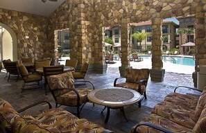 An outdoor lounge area with stone pillars, wicker furniture, and a pool in the background.