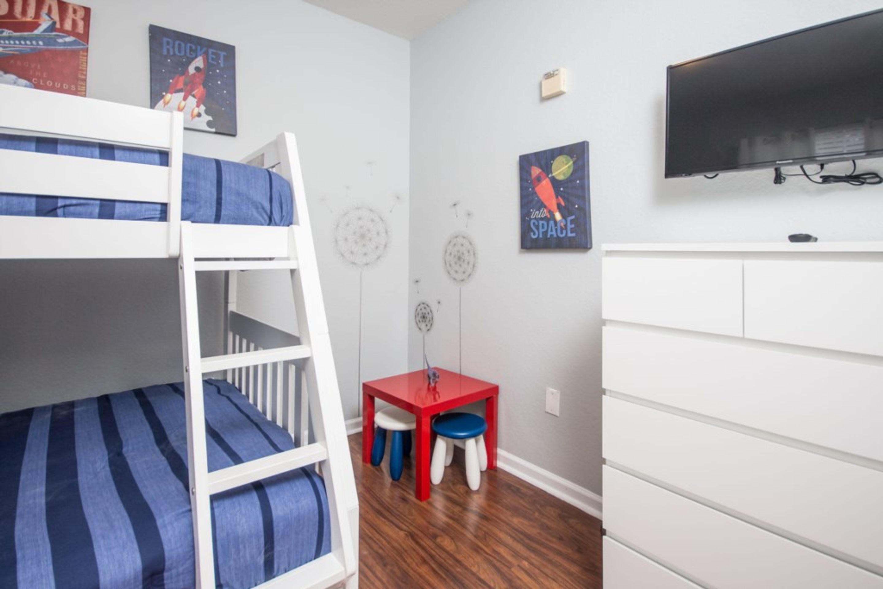 Een kinderbed met stapelbedden, een blauw gestreepte matras, een kleine rode tafel met krukjes en een witte dressoir met een flatscreen televisie erboven.