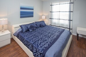 A bedroom with a large bed, two bedside tables, a nightstand, a window with curtains, and a wall-mounted artwork. - Windsor Palms 2300 - 101 (Kissimmee)