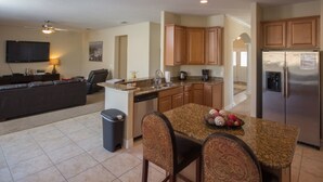 A modern kitchen with granite countertops, wooden cabinets, a stainless steel refrigerator, and a dining area with patterned chairs. - The Shire at West Haven 841 (Davenport)