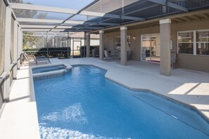 Une piscine intérieure protégée par une paroi de verre, une terrasse ensoleillée avec des sièges et une vue sur l’extérieur.