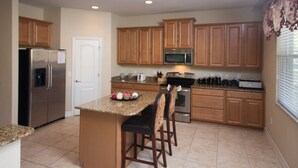 A kitchen with wooden cabinets, a stainless steel refrigerator, a microwave, and a stove. There are two bar stools and a small island with a granite countertop. - The Shire at West Haven 841 (Davenport)