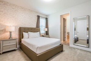 Quarto com parede de tijolos, uma cama grande, duas mesas de cabeceira com luminárias, um espelho e uma porta que dá para outro cômodo.