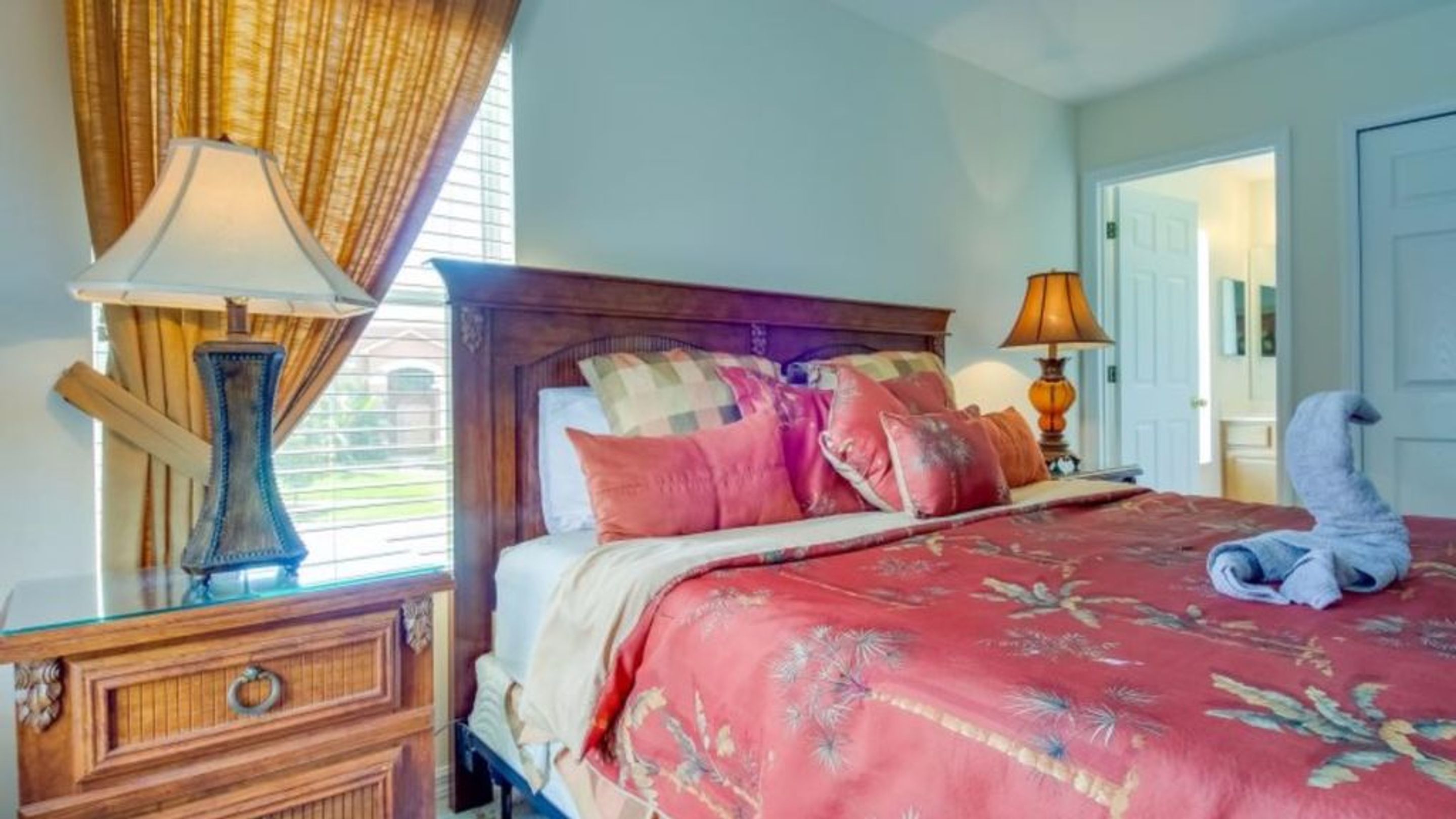 A bedroom with a wooden bed, multiple pillows, a nightstand with a lamp, and a window with curtains.
