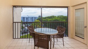 A balcony with a glass table and four chairs, a door with a window, and a view of buildings and trees. - Fort Myers Beach Area 7327 - 409 (Fort Myers Beach)