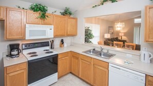 A kitchen with wooden cabinets, a microwave, stove, sink, and dishwasher. There are plants on the cabinets and a dining area visible in the background. - Westridge 110 (Davenport)
