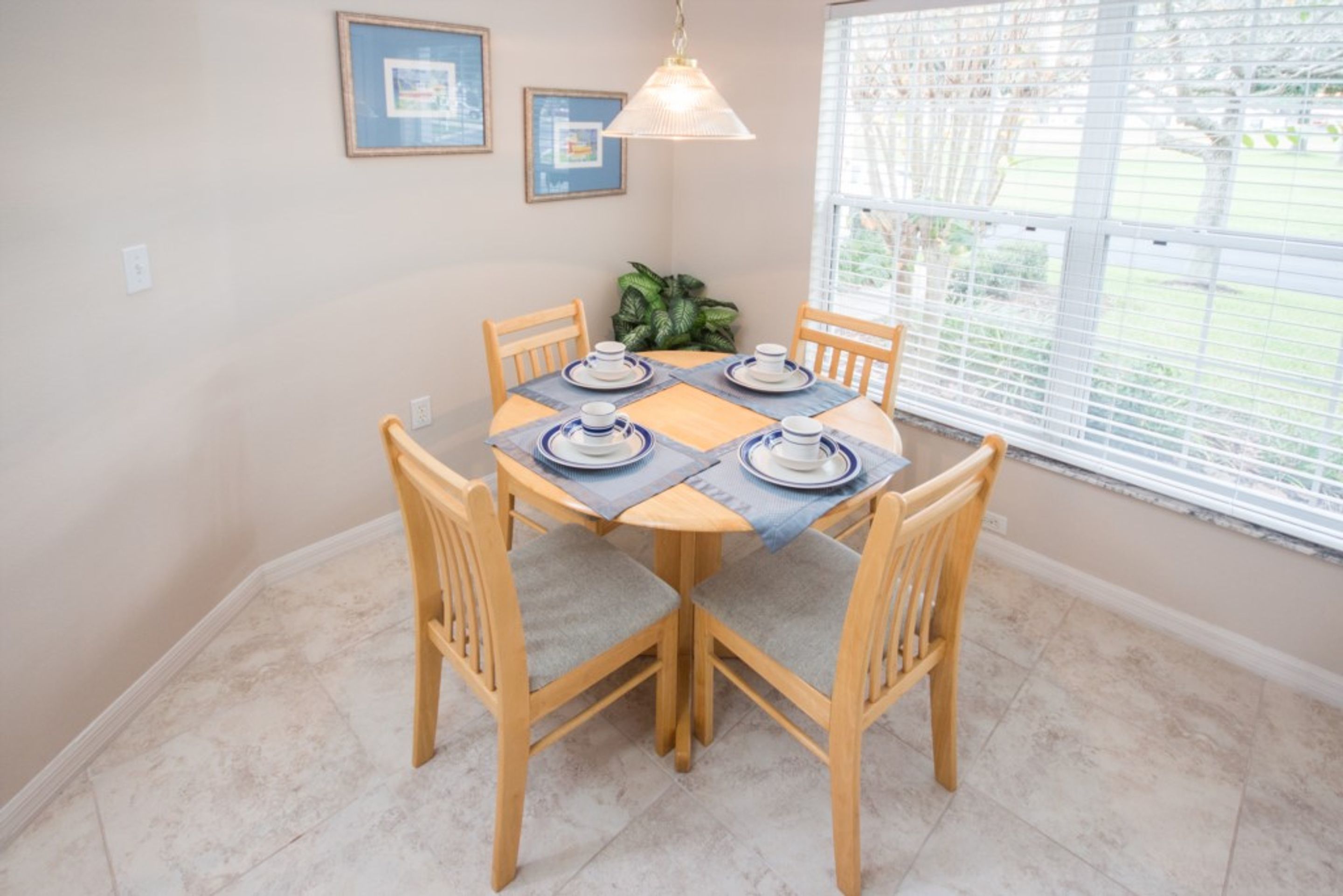 Un coin repas avec une table dressée pour quatre personnes, entourée de chaises en bois, d’un luminaire suspendu et d’une fenêtre avec des stores.