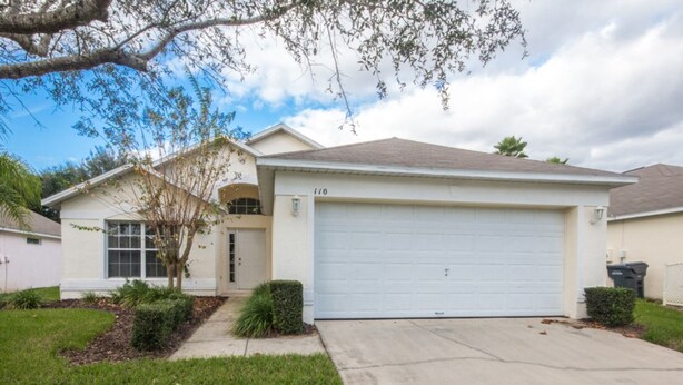 A single-story house with a white garage door, a small front yard with grass and a few bushes, and a tree with sparse leaves. - Westridge 110 (Davenport)