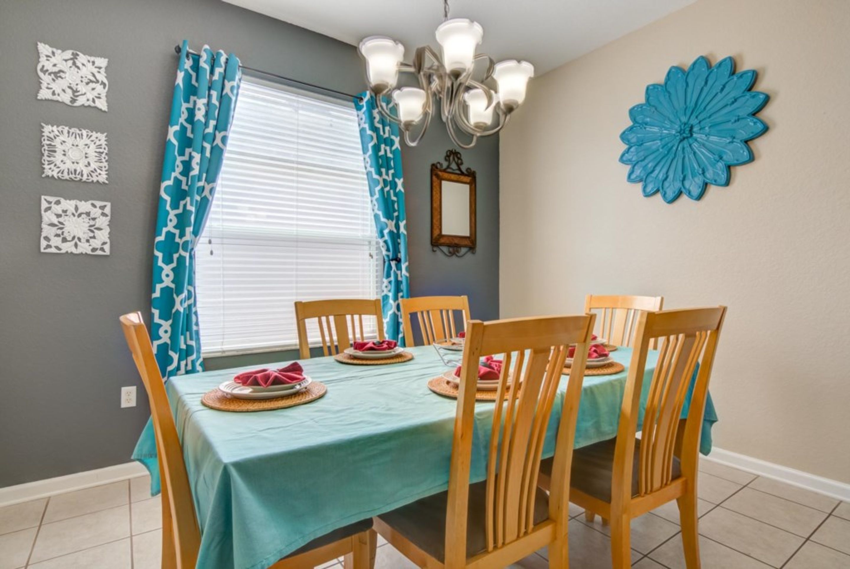 A dining area with a table set for four, wooden chairs, blue curtains, and a decorative blue flower on the wall.