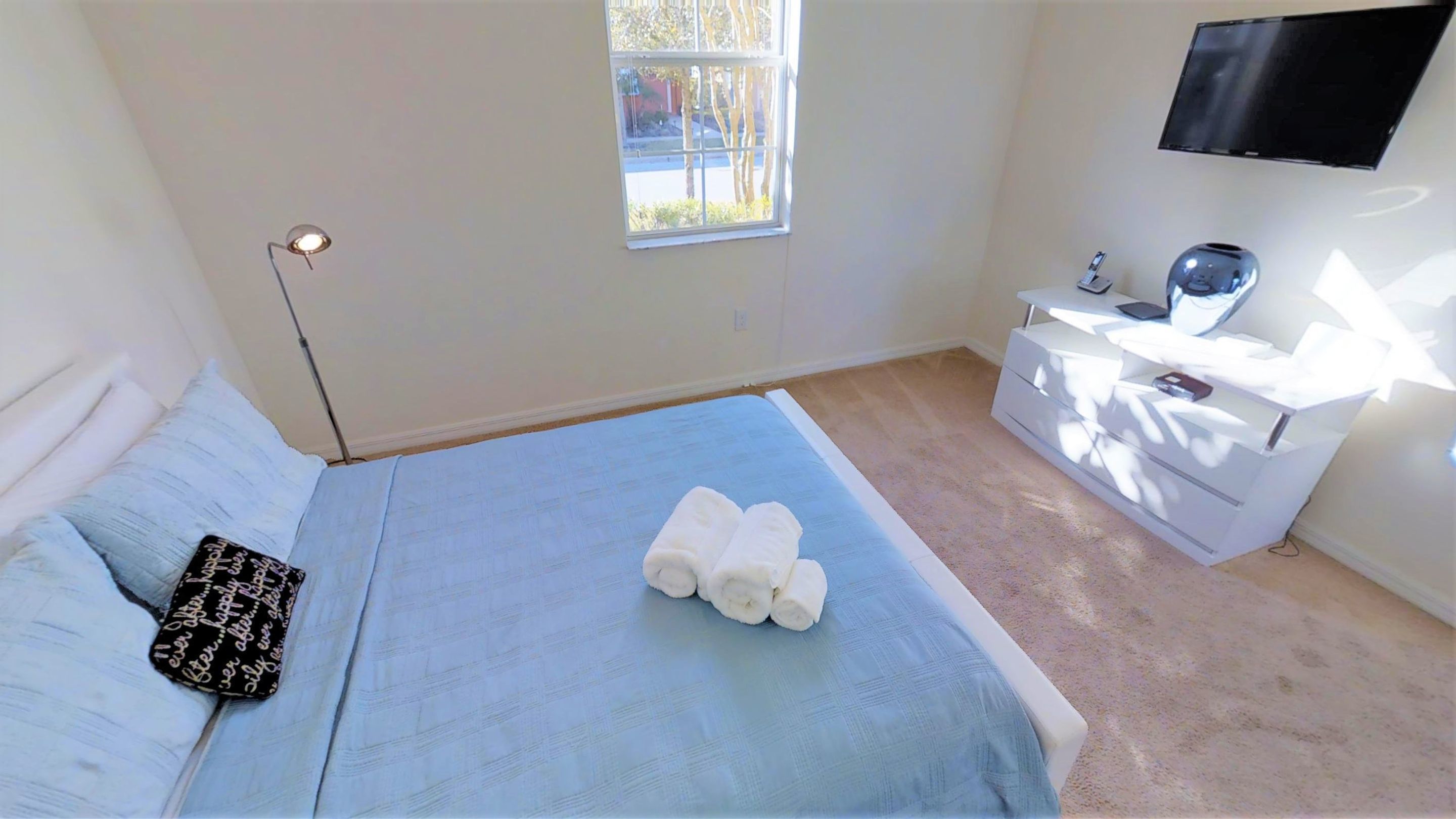 Een slaapkamer met een bed, een nachtkastje met een lamp, een aan de muur bevestigde televisie, en een raam met uitzicht.