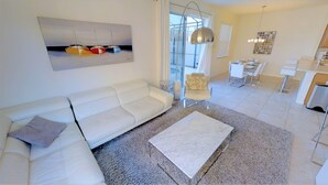 A modern living room with a white sofa, a coffee table, and a painting of boats. There is a dining area with a table and chairs, and a kitchen area with a sink and countertop. - Bella Vida 541 A (Kissimmee)