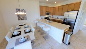 A modern kitchen with a central island, wooden cabinets, stainless steel appliances, and a dining area with white chairs and a table set for four. - Bella Vida 541 A (Kissimmee)