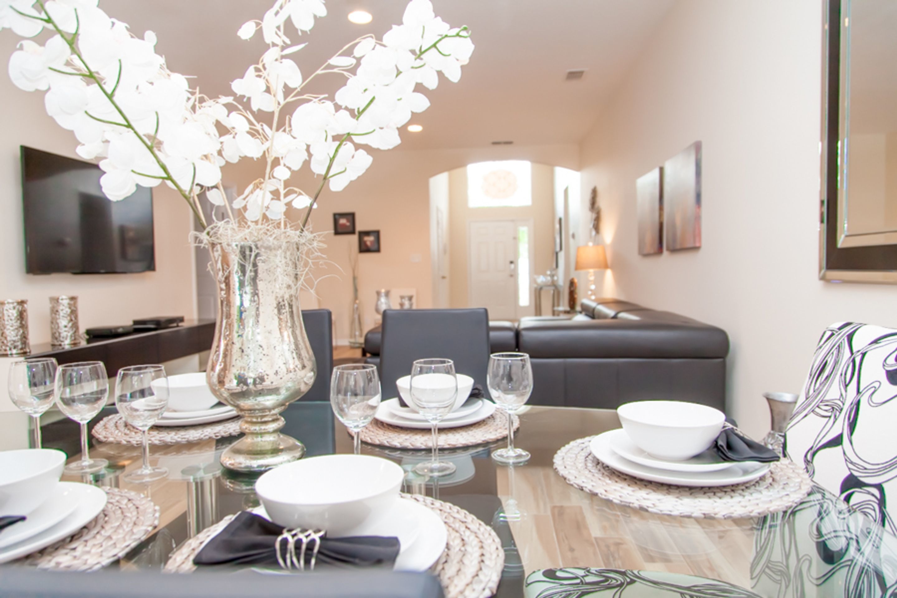 A dining area with a glass table set for four, white plates, bowls, and wine glasses. There is a decorative vase with white flowers on the table.