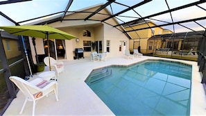 A covered outdoor swimming pool with lounge chairs and a table, surrounded by a glass enclosure. - Seasons 1175 (Kissimmee)