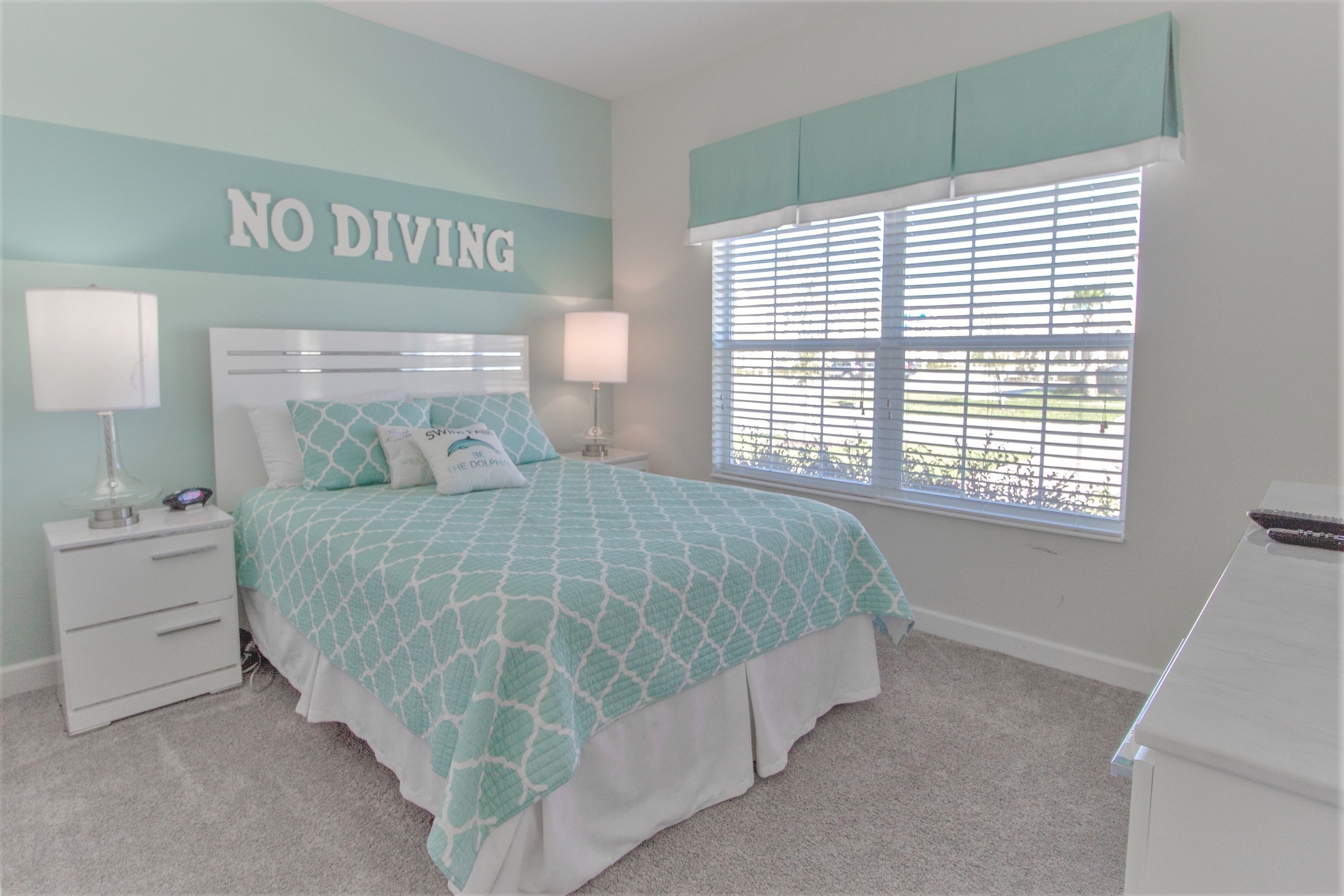 Ein Schlafzimmer mit Bett, Nachttischen, einem Fenster mit Jalousien und einem Wandschild mit der Aufschrift 'NO DIVING'.