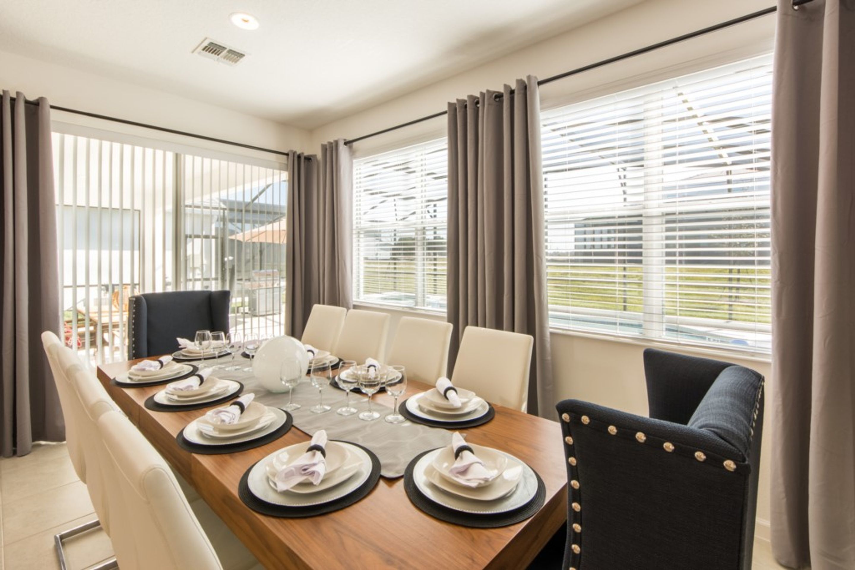 A dining area with a wooden table set for six, surrounded by chairs, with large windows and curtains.