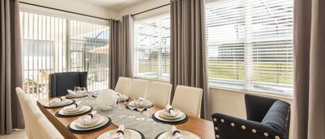 A dining area with a wooden table set for six, surrounded by chairs, with large windows and curtains.