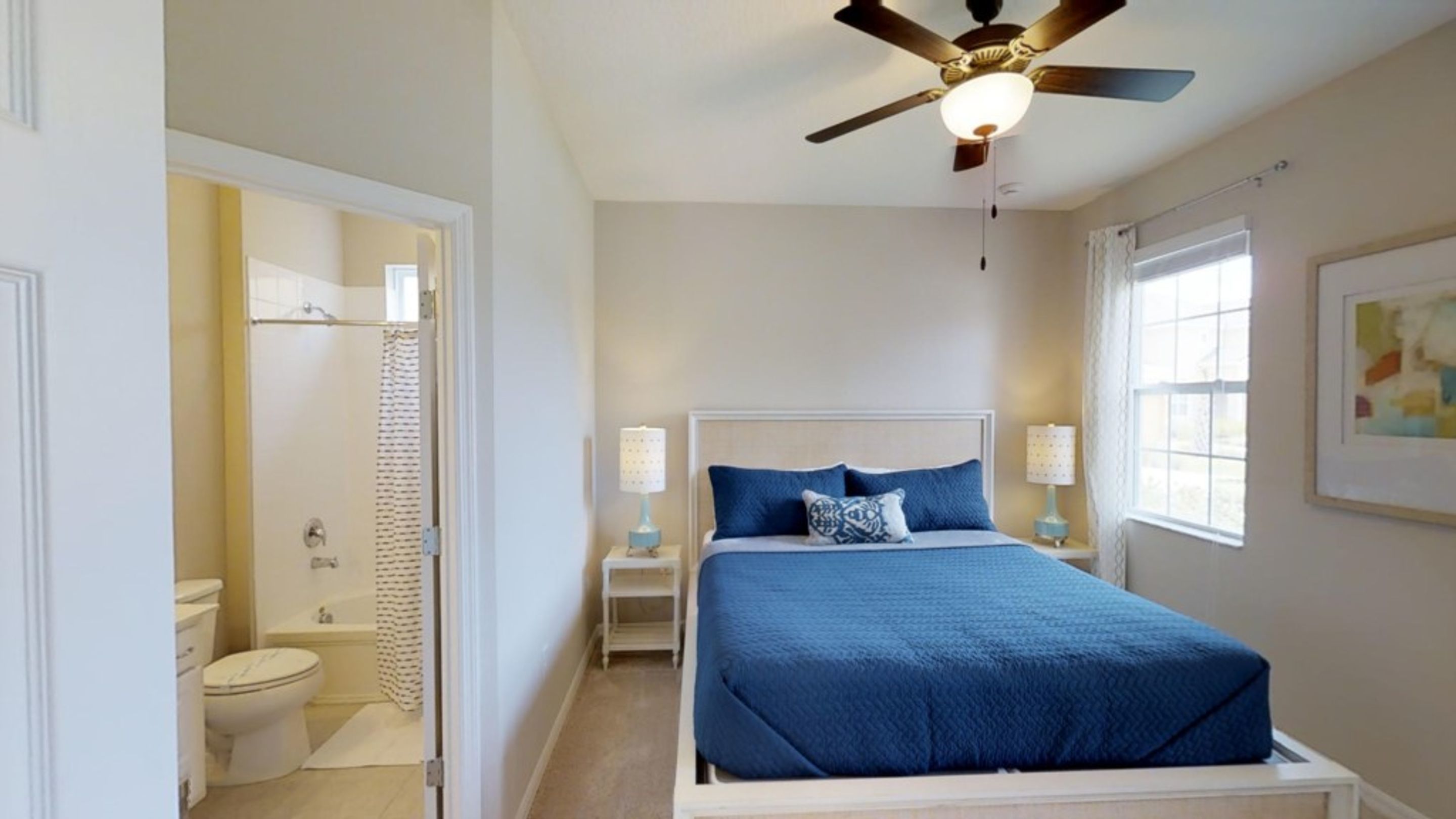 A bedroom with a bed, two bedside tables, a ceiling fan, a window, and a bathroom visible through an open door.