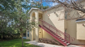A two-story building with a red staircase leading to the second floor. There is a small front yard with a tree and a sidewalk. - Club Cortile 2730a (Kissimmee)