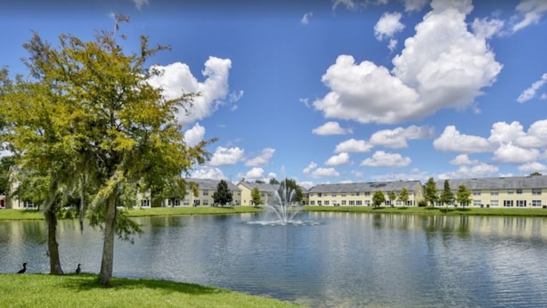 A serene lake with a fountain, surrounded by well-maintained grass and trees, with buildings in the background. - Club Cortile 2730a (Kissimmee)