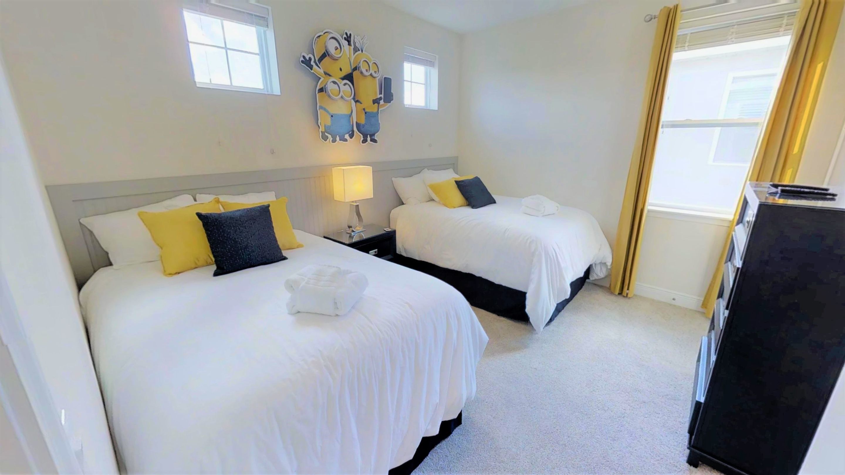 A hotel room with two beds, a nightstand, a lamp, and a Minion decoration on the wall.