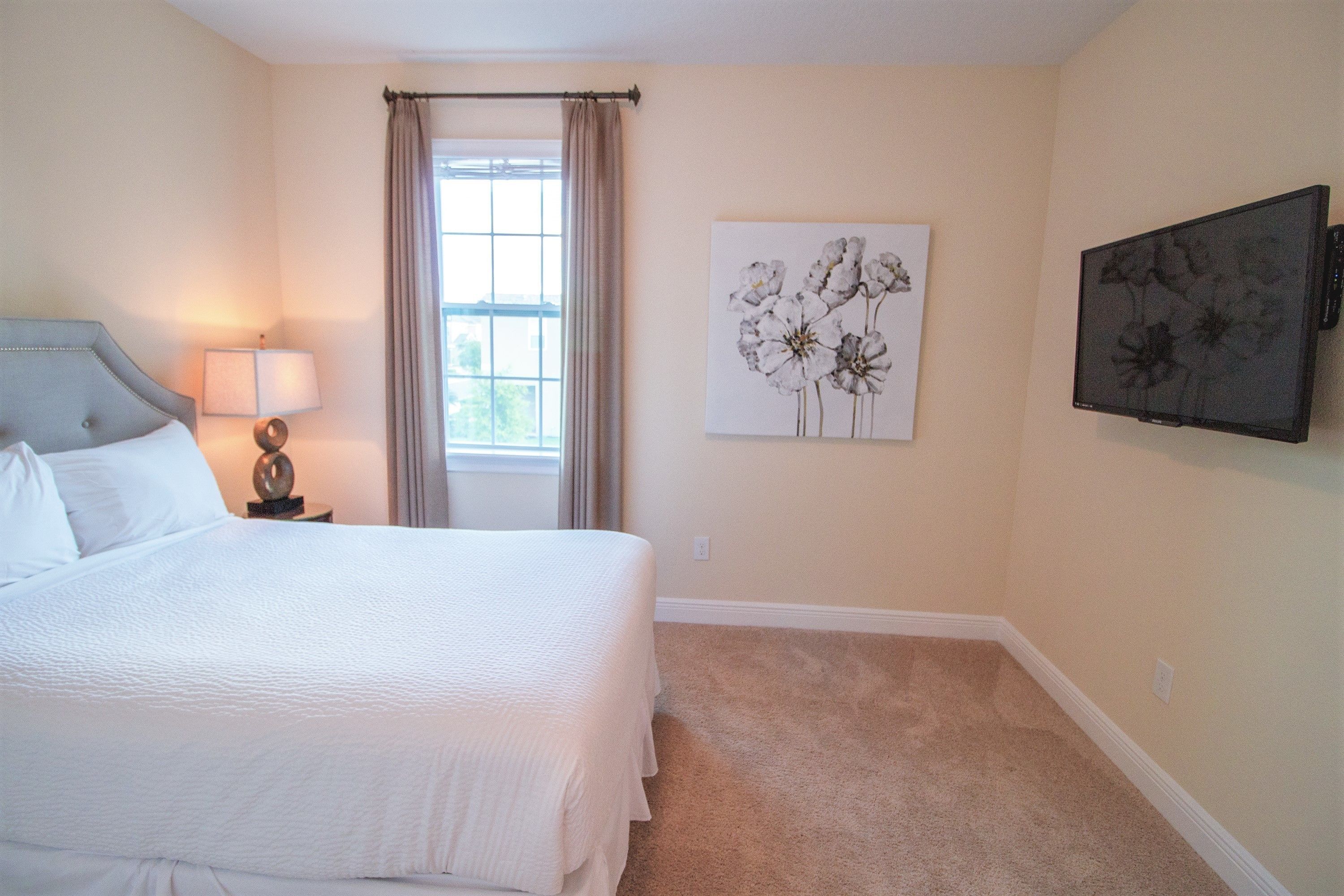 A bedroom with a bed, a nightstand, a lamp, a window with curtains, and two wall-mounted artworks.
