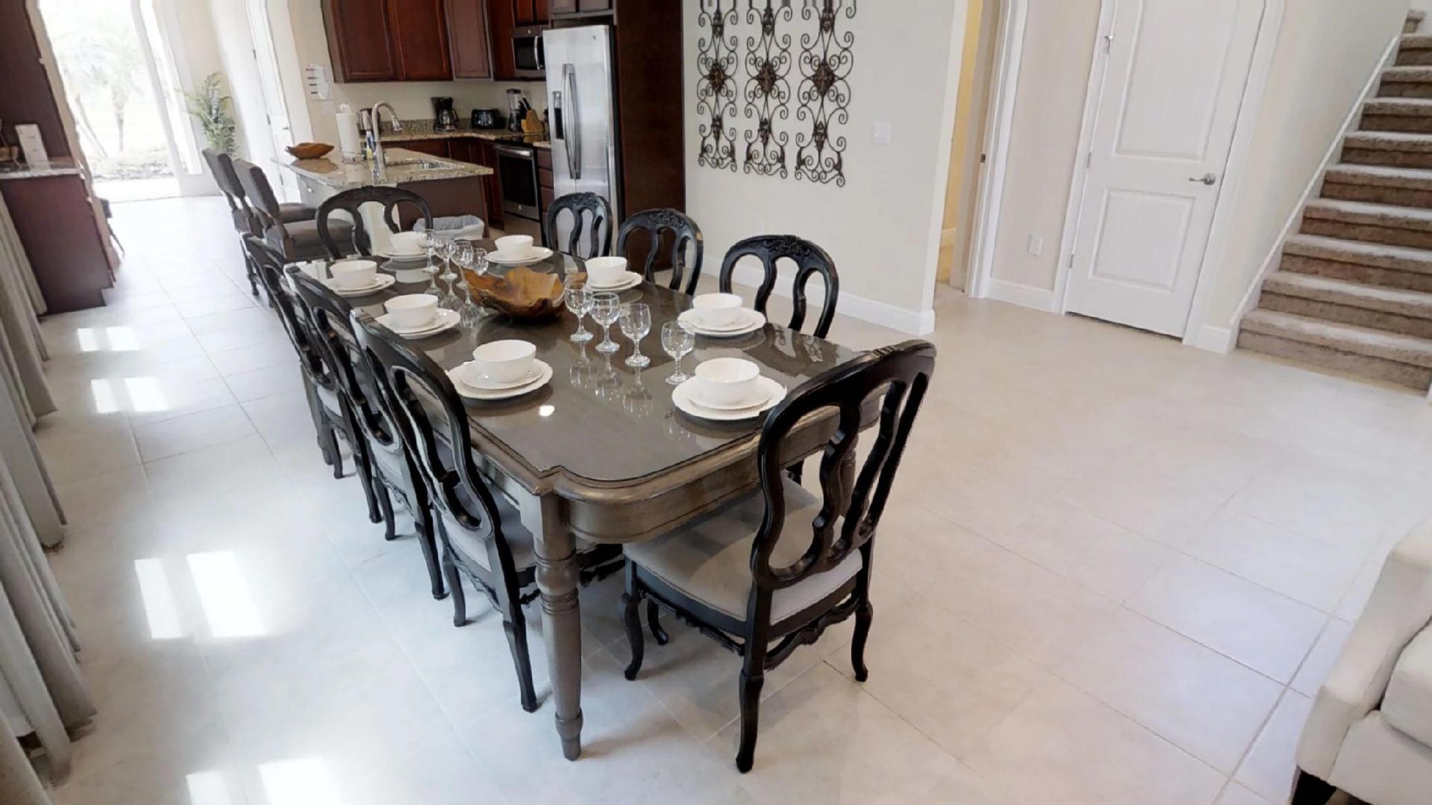 On aperçoit un coin repas avec une longue table dressée pour six personnes, entourée de chaises. Au fond, on distingue une cuisine avec des meubles en bois foncé et un réfrigérateur.